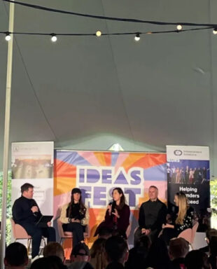 Five speakers sitting in chairs on stage under a canvas canopy giving a talk to an audience